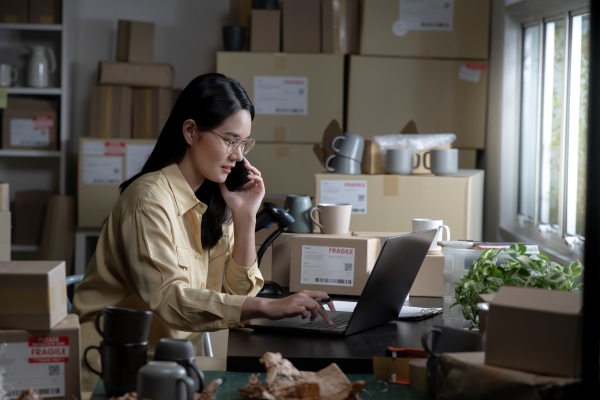 asian-woman-online-seller-confirming-orders-from-customer-phone-laptop-ecommerce-business-owner-using-phone-looking-laptop-store-warehouse